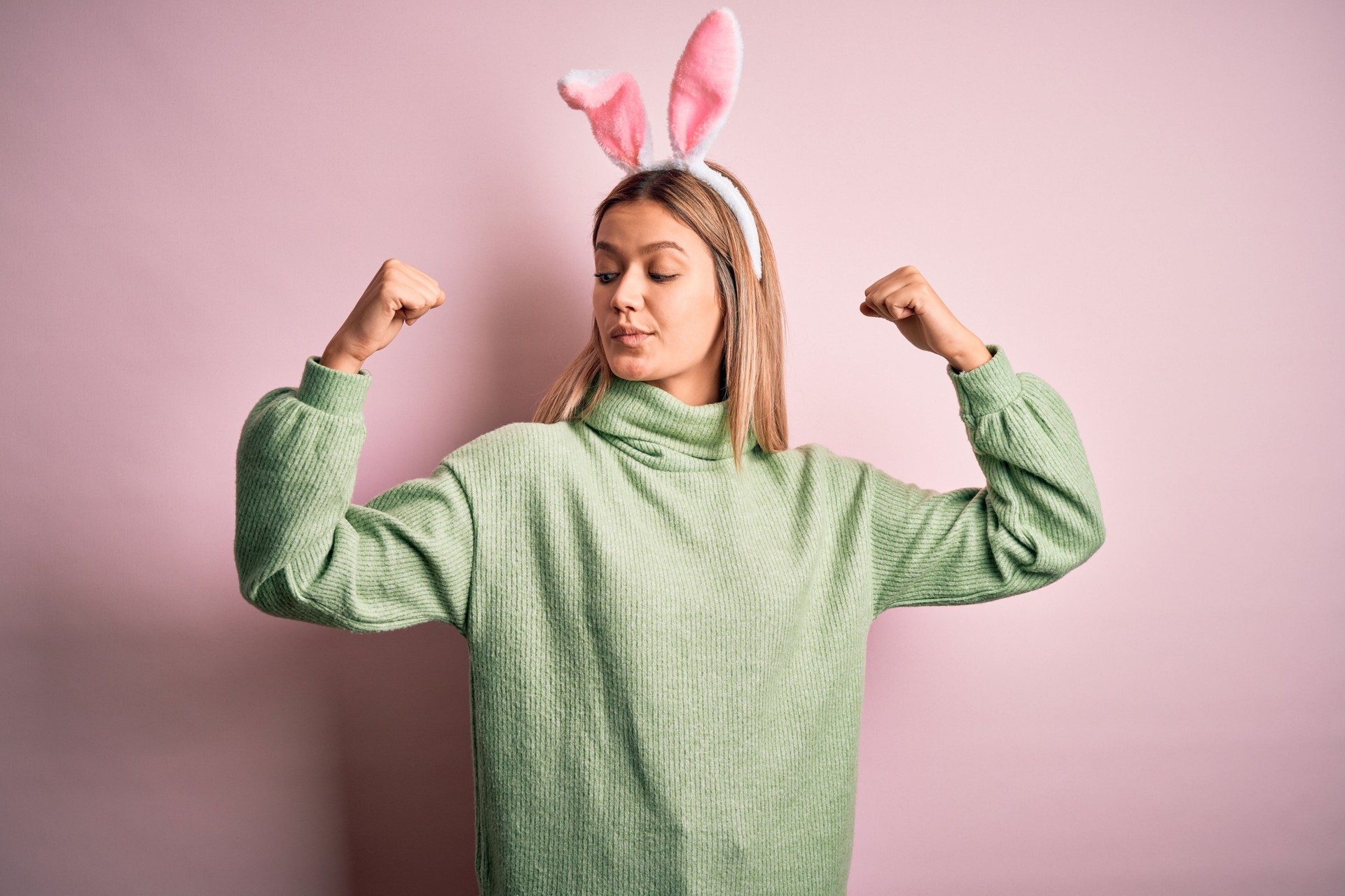 Young beautiful woman wearing easter rabbit ears standing over isolated pink background showing arms muscles smiling proud. Fitness concept.